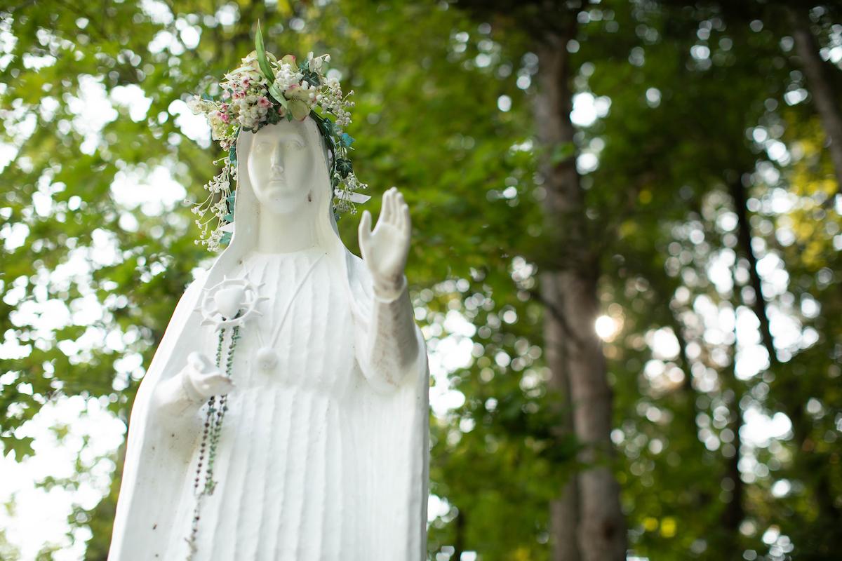 Statue of our lady fatima with a crown of flowers and rosary and a backdrop of leaves